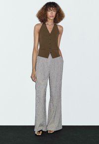 Gilet marrone senza maniche con scollo a V e cinque bottoni, abbinato a pantaloni larghi grigi chiari con texture. Sandali semplici completano l'outfit.