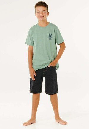 Ragazzo in piedi a piedi nudi, indossa una maglietta verde con grafica e pantaloncini neri fino al ginocchio, sorride con una mano in tasca su sfondo bianco.