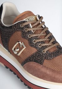 Sneaker marrone e nera con rete, suede e accenti metallici. Caratterizza motivi testurizzati, hardware dorato e suola bianca spessa con dettagli rossi.