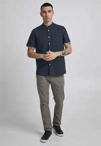 Cămașă cu mânecă scurtă, cu nasturi, dintr-un material navy închis, cu guler în picioare, un buzunar pe piept și un finisaj texturat. Purtați-o cu pantaloni gri deschis.