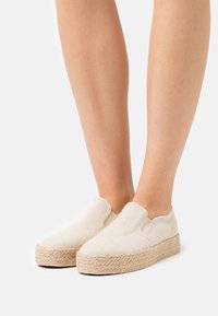 Scarpe slip-on in tessuto naturale di colore crema, con una suola in juta tessuta texture e una punta arrotondata.