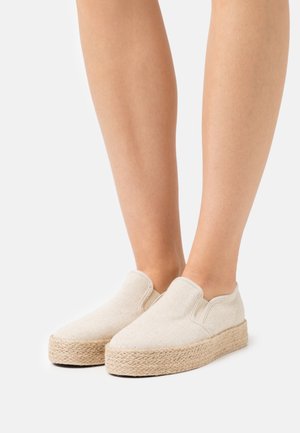 Zapatos slip-on de tela natural en color crema, con una suela de yute tejido texturizado y un diseño de punta redondeada.