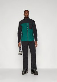 Zwarte en groene fleecejas met ritssluiting aan de voorkant en zakken, gecombineerd met zwarte broek en skischoenen; houdt een skibril vast.