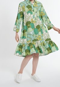 Grön blommig klänning med krage, långa ärmar och en rynkad volangkant, tillverkad av lättviktigt tyg. Bärs med vita sneakers.