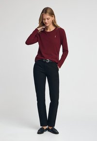 Bordeauxrotes Langarmshirt mit rundem Ausschnitt, kombiniert mit schwarzer Hose und schwarzen Schuhen, mit einem dezenten, auf dem Shirt gestickten Logo.