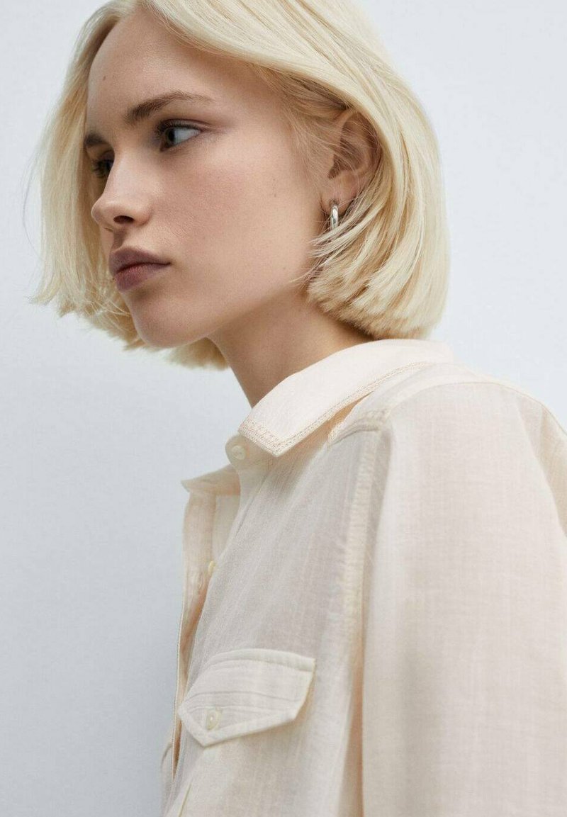 Capelli biondi, primo piano di una persona che indossa una camicia button-up beige chiaro con colletto e tasca, mostrando una trama liscia.