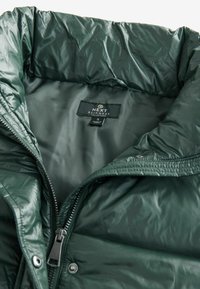 Zelená, lesklá nafukovacia bunda s vysokým golierom. Obsahuje zips a nálepku s nápisom "NEXT RAINWEAR COLLECTION." Hladká textúra.