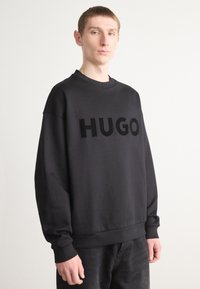 Black sweatshirt featuring bold, raised "HUGO" logo on the front. Made from soft fabric, it has a ribbed neckline and cuffs.