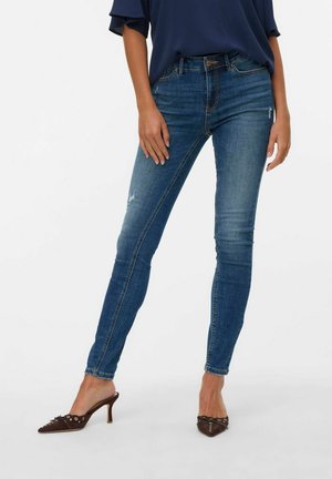 Skinny blue denim jeans with a faded finish, slight distressing, and a mid-rise waist, paired with dark brown heeled mules.