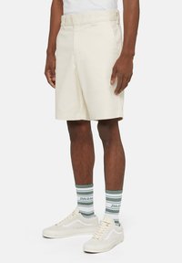 Pantaloni beige con frontale piatto e passanti per cintura, fino al ginocchio. Indossati con calzini a righe verdi e bianchi e sneakers bianche.