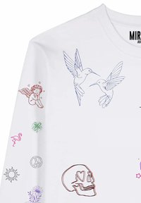 Chemise blanche à manches longues avec des dessins au trait de deux colibris sur la poitrine et divers petits symboles tels qu'un crâne, un ange, un flamant rose et un signe de paix sur la manche.