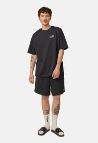 Czarny bawełniany t-shirt z krótkim rękawem i logo, zestawiony z czarnymi szortami. Noszony z białymi skarpetkami i ciemnymi sandałami. Widoczne tatuaże na ramionach.