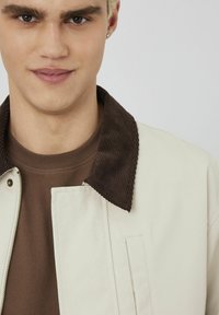 Veste beige clair avec un col en velours côtelé marron foncé ; équipée d'une fermeture éclair à l'avant et d'une poche rectangulaire sur la poitrine. Chemise marron en dessous.