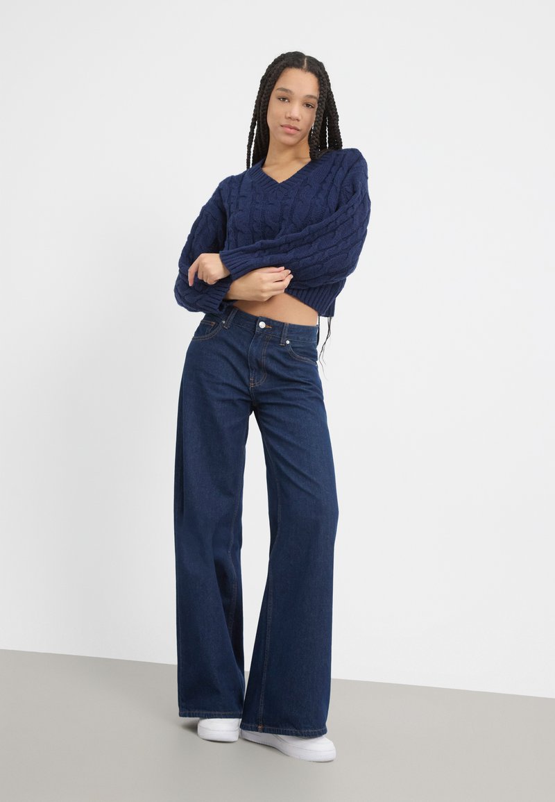 Pull en maille torsadée marine avec un col en V et une coupe courte, associé à un jean en denim foncé à jambes larges et des baskets blanches, sur fond uni.