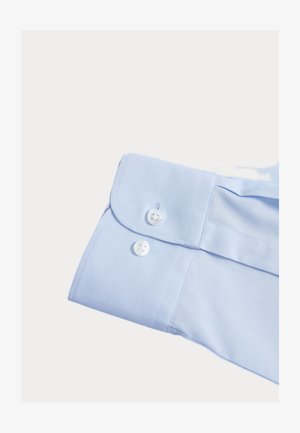 Puño de camisa azul claro hecho de tela suave, con dos botones blancos y un diseño de borde redondeado. No se observan patrones ni adornos.