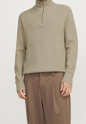 Beige ribstrikket pullover-trøje med kvart lynlåskrave båret med brune plisserede bukser.