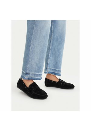 Person wearing light blue cropped jeans and black suede loafers with lace details, standing on a neutral background.