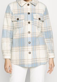 Geruite shirtjack in zachte, fluffy stof met blauwe, witte en beige kleuren. Beschikt over twee borstzakken en donkere knopen.