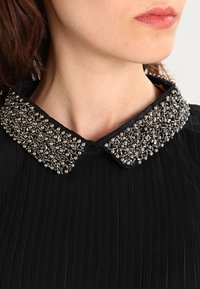 Svart topp med en plisserad design som har en pärlad, metallisk krage. Kragen är prydd med silverpärlor i olika storlekar.