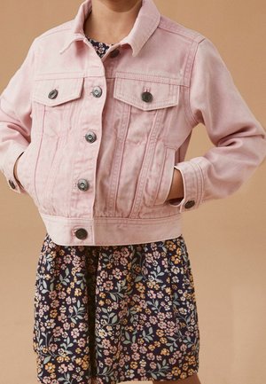 Bambino che indossa una giacca di jeans rosa chiaro con bottoni, con le mani nelle tasche e un vestito scuro a fiori sotto.