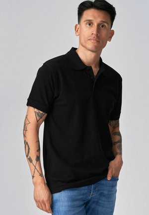 Muž s tetovaniami na rukách, oblečený v čiernom polokošeli a modrých džínsoch, stojaci pred jednoduchým sivým pozadím.