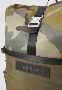Kamuflasjet bag laget av slitesterkt stoff med rulleslutt, justerbare svarte stropper og en "REPLAY"-lapp foran.