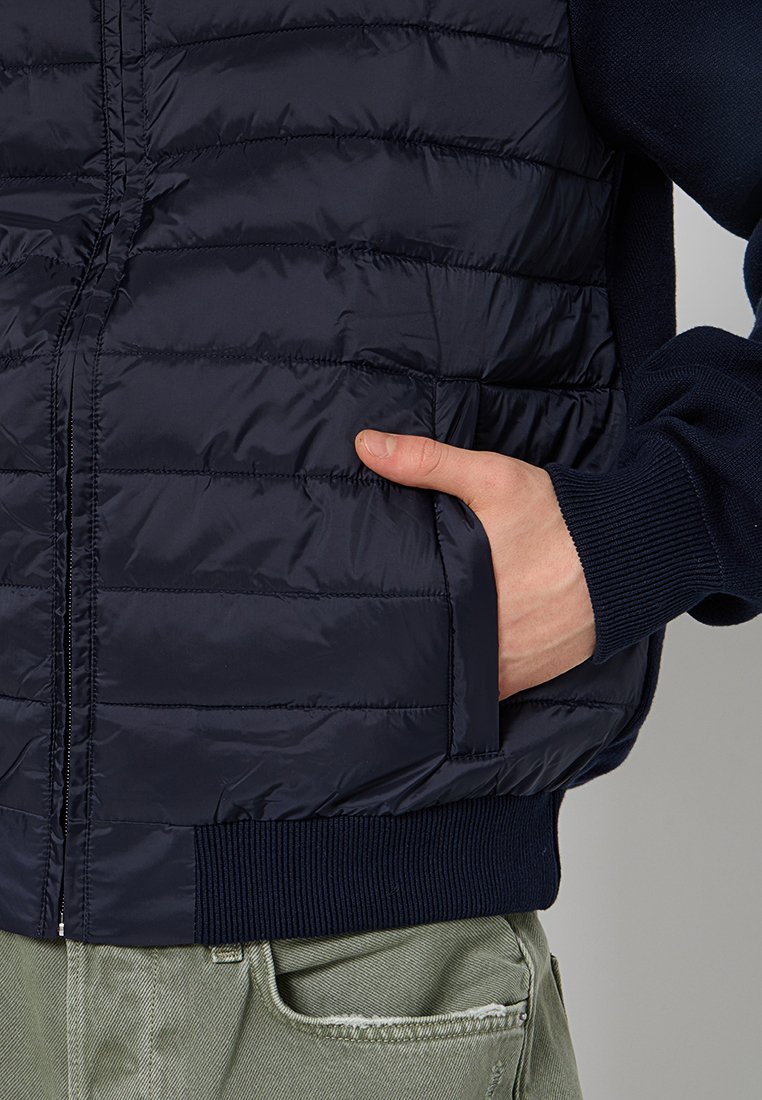 Marineblauer, dicker Weste mit horizontaler Naht, Reißverschluss und gerippten, marineblauen Bündchen. Verfügt über seitliche Taschen und eine glatte, glänzende Textur.