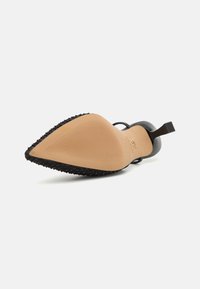 Svarte høyhælte sko med spiss tå, med en teksturert svart overdel og en glatt beige såle med ribbet detalj. Firkantet helldesign.