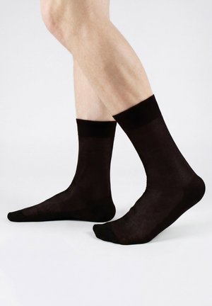 Beine, die durchsichtige schwarze Kniestrümpfe tragen, stehen auf einer weißen Oberfläche mit einem leicht gehobenen Fuß.