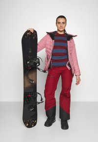 Snøbrett med mørk topp og lys bunn, holdt av en person i en rosa isolert jakke, stripete blå og maroon skjorte, og røde bukser.