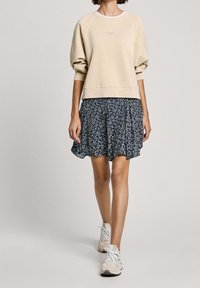 Felpa beige con polsini a coste sopra una gonna plissettata scura a fiori, abbinata a sneakers di colore chiaro. Tessuto morbido con vestibilità rilassata.