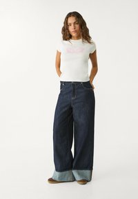 Biała koszulka z krótkim rękawem z różowym wzorem graficznym, w połączeniu z szerokimi, ciemnoniebieskimi dżinsami z jasnoniebieskim detalem na brzegach oraz brązowymi butami.