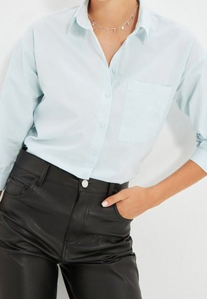 Frau trägt ein hellblaues, geknöpftes Hemd mit Brusttasche, schwarze, hochtaillierte Lederhose und eine zarte silberne Halskette, Hand in der Tasche.