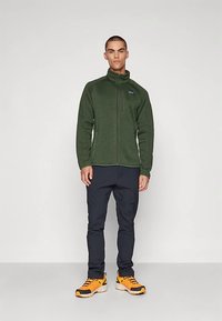 Moški stoji ob beli podlagi, oblečen v zeleni zadrgo jopič iz flisa, črne hlače in svetlo oranžne pohodne čevlje.