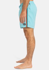 Shorts de bain bleu clair avec une ceinture élastique, poches latérales et une bande orange. Dotés d'un écusson noir sur l'ourlet.