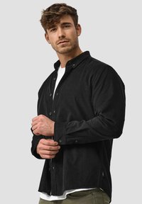Camisa negra de pana con botones y mangas largas, tejido texturizado y camiseta interior blanca de contraste, mostrada con un modelo en un entorno neutro.