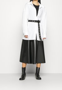 Weißes, übergroßes Hemd mit langen Ärmeln, schwarzer, lederner A-Linien-Rock und schwarzen Stiefeletten. Das Hemd hat einen kontrastierenden schwarzen Gürtel.