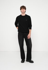 Fekete kötött pulóver kerek nyakkal, sötét farmerrel és fekete cipővel párosítva. A modell göndör hajú, egyszínű háttér előtt áll.