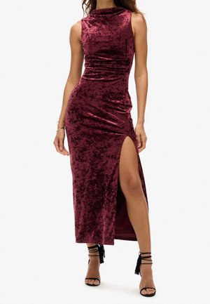 Robe midi bordeaux en velours sans manches avec une fente haute sur un côté, portée avec des sandales à talons hauts noires à lanières et des bracelets à un poignet.