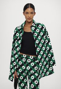 Giacca a fantasia verde e nera con fiori bianchi, abbinata a pantaloni a fantasia coordinati e una maglietta nera. Tessuto sintetico liscio.