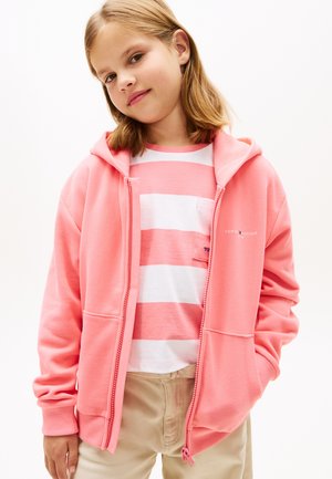 Mladé dievča má na sebe ružovú mikinu na zips cez ružovo-biele pruhované tričko a béžové nohavice, stojí s jednou rukou v vrecku.