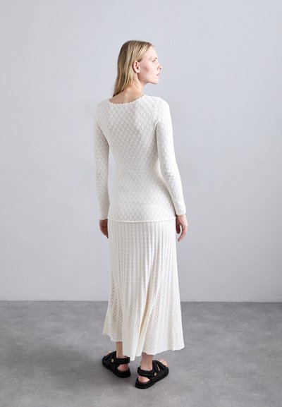 Witte gebreide top met lange mouwen en ruitpatroon, gecombineerd met een bijpassende geplooide maxi rok. Zwarte sandalen maken de outfit compleet.