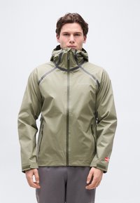 Νεαρός άνδρας που φοράει ελαφρύ ανοιχτοπράσινο αδιάβροχο της Columbia με κουκούλα και γκρι παντελόνι, στέκεται μπροστά σε απλό φωτεινό φόντο.