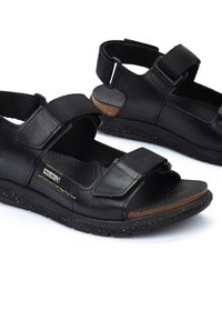 Las sandalias de cuero negro cuentan con correas ajustables de Velcro, una plantilla perforada y una suela texturizada con un diseño moteado.