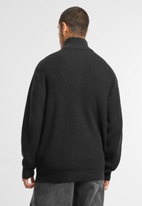 Pull-over en maille noir à côtes avec col montant et manches longues, présentant une coupe décontractée et un tissu texturé.
