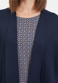 Mörkblå stickad kcardigan över en mönstrad blus med blå, brun och krämfärgad färg, med rund hals och geometrisk design.