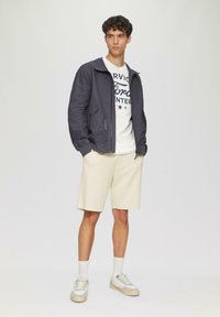 Chaqueta bomber navy con cremallera frontal, pantalones cortos beige, camiseta blanca con gráficos azules y zapatillas ligeras. Tejidos suaves, ajuste casual.