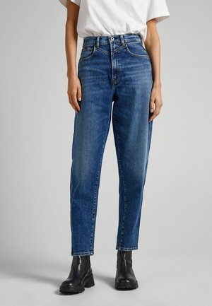Person trägt hochtaillierte blaue Jeans und schwarze Knöchelstiefel und steht vor einem einfarbigen Hintergrund.