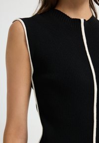 Canottiera nera senza maniche a costine con strisce verticali bianche lungo le aperture delle braccia e il centro davanti, indossata da una persona con pelle chiara.