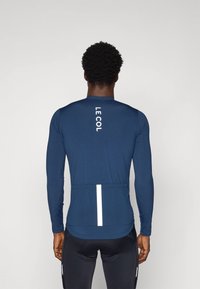 Maillot de cyclisme à manches longues bleu marine avec des accents réfléchissants, présentant un logo au dos, en matériau qui évacue l'humidité et au design ajusté.